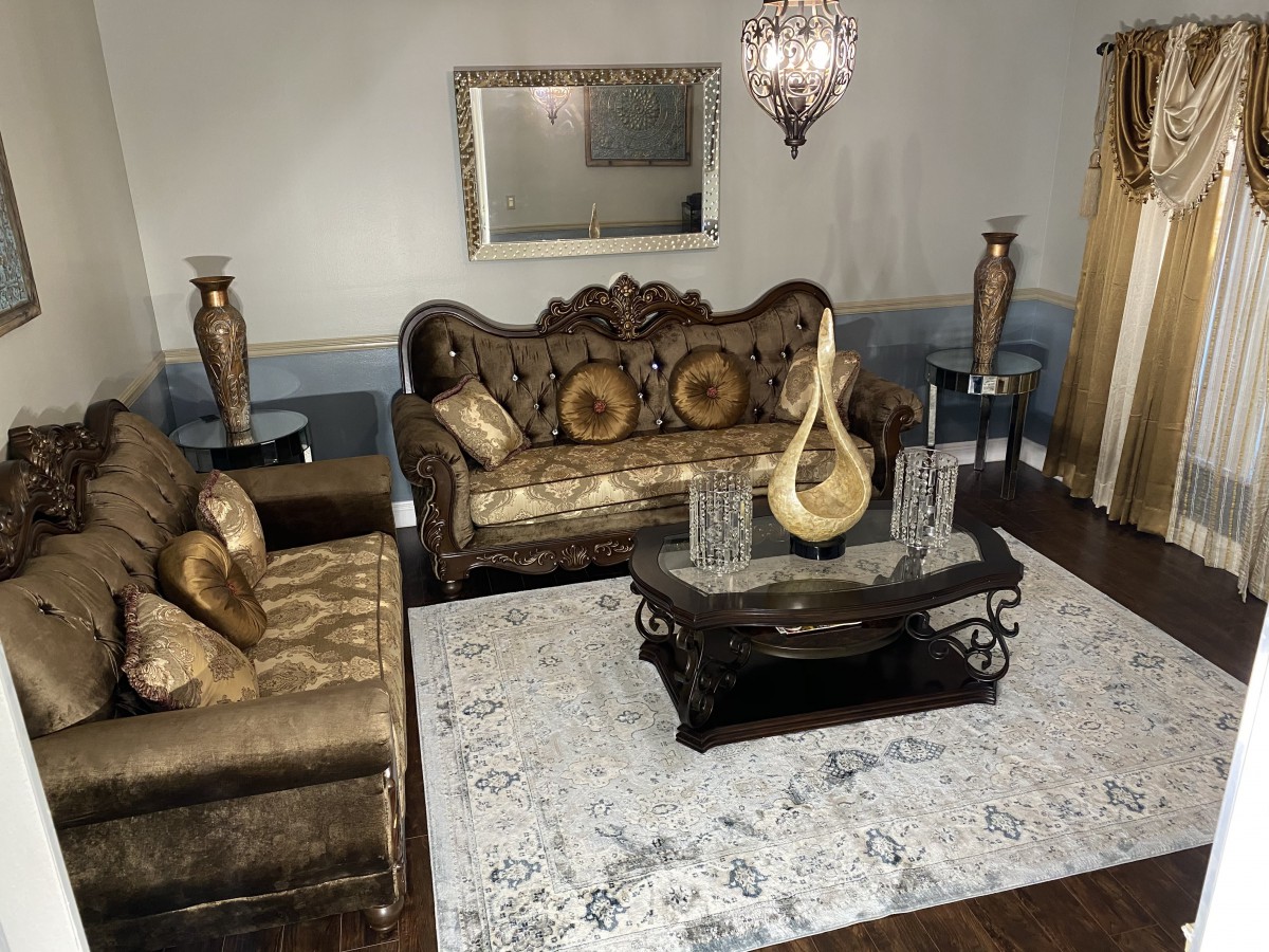 A traditionally decorated living room featuring an ornate sofa set, a central coffee table with decorative pieces, and elegant vases on either side. A large mirror hangs above the sofa, complemented by a chandelier.