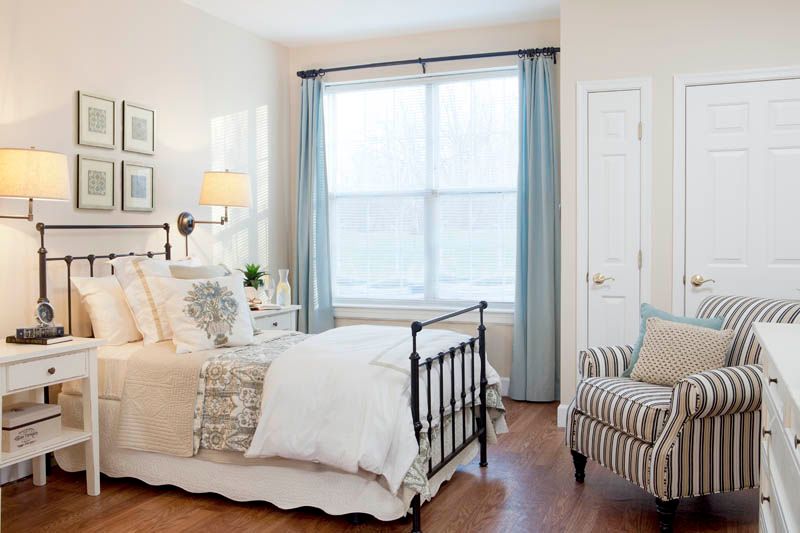 A cozy and bright bedroom featuring a neatly made bed, soft lighting from a bedside lamp, and large windows with pale blue curtains.