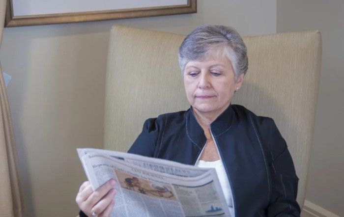 A woman with gray hair reads a newspaper while sitting in a chair.