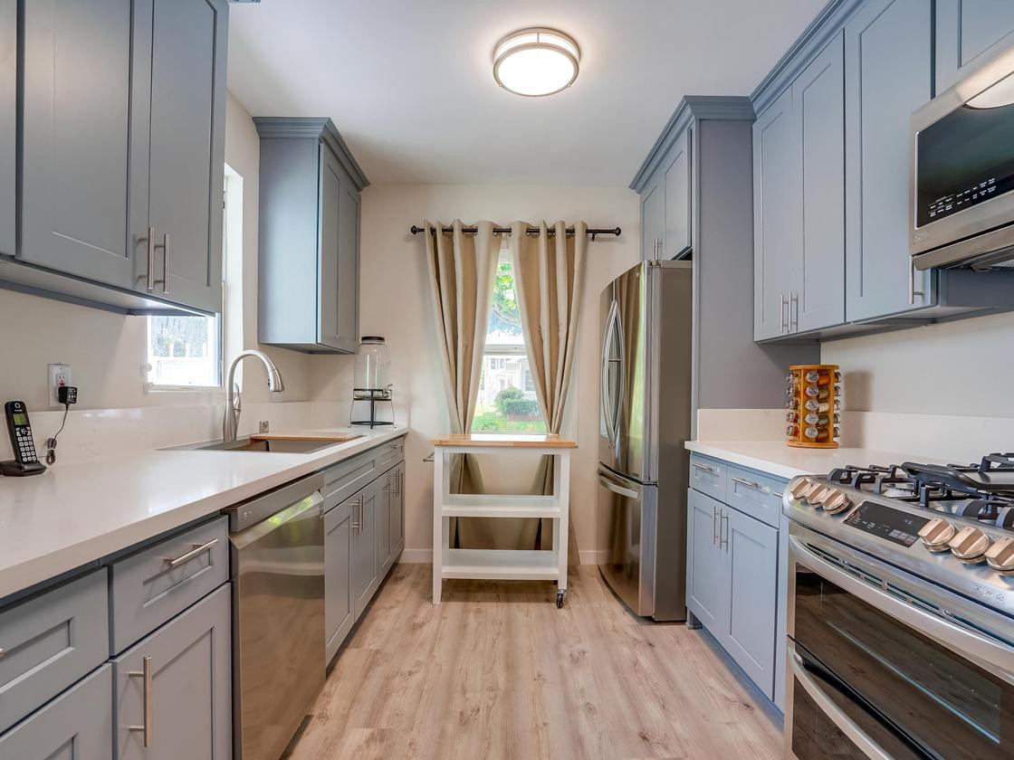A modern kitchen with gray cabinets, stainless steel appliances, and wooden flooring, illuminated by natural light.