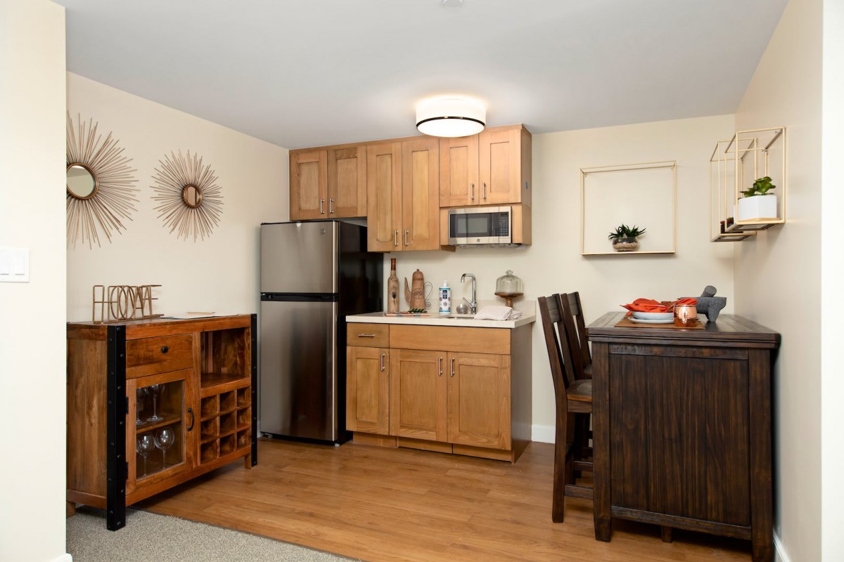 A modern kitchen interior with wooden cabinets, featuring a refrigerator, microwave, and dining area.