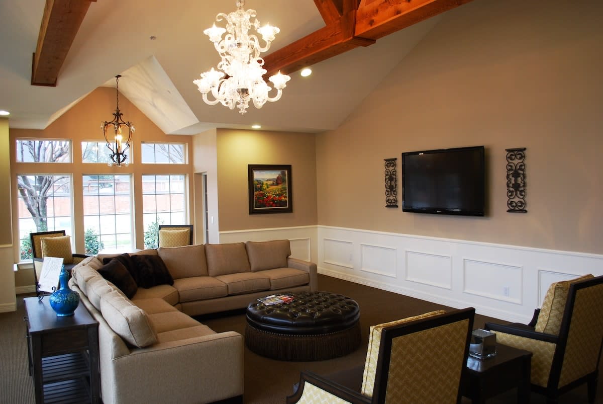 A stylish living room featuring a modern chandelier, large sofa, and wall-mounted TV with exposed wooden beams on the ceiling.