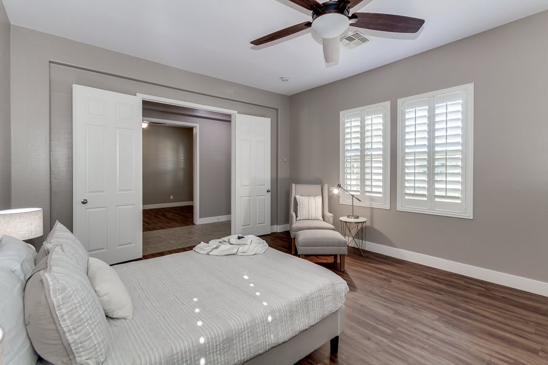 A modern bedroom with neutral tones, featuring a bed, chair, and ceiling fan, illuminated by natural light through shuttered windows.