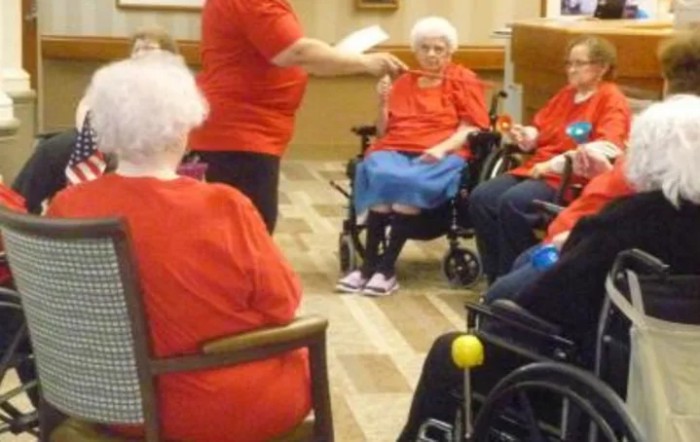 A group of elderly people wearing red participate in an indoor activity, some seated in wheelchairs.