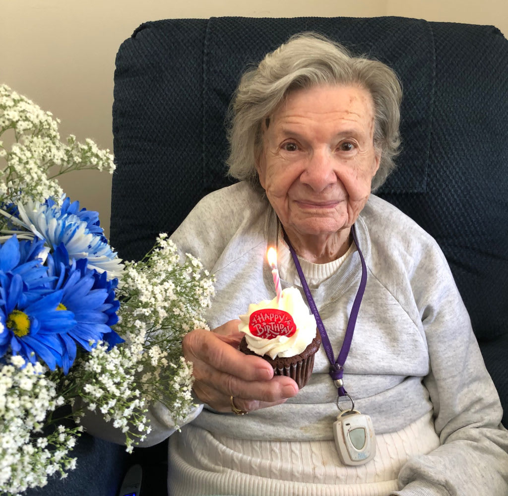 An elderly woman holding a cupcake with a candle, sitting next to a bouquet of blue flowers.