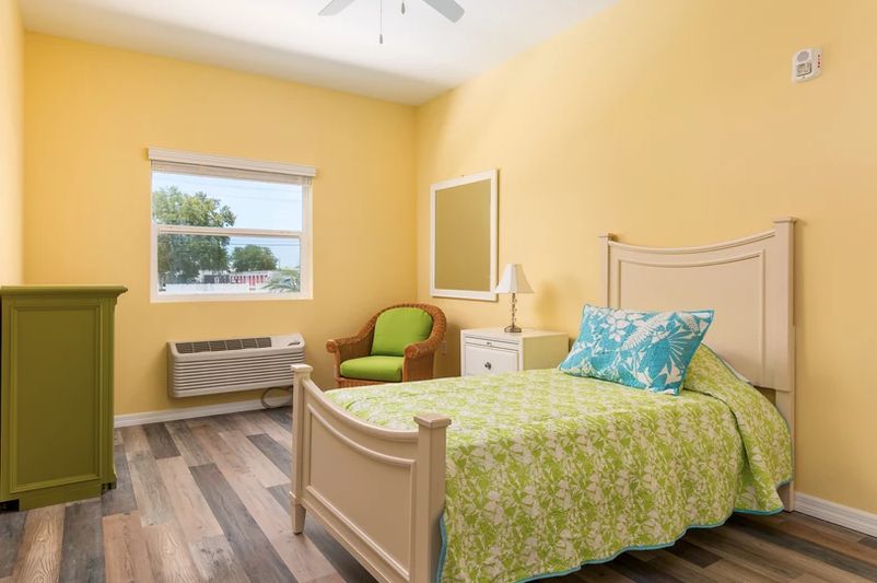 A cozy bedroom with bright yellow walls featuring a single bed, an armchair, and wood flooring.