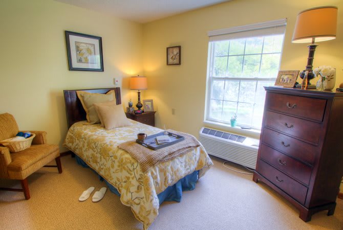 A cozy bedroom with a single bed, warm lighting from a lamp, and a large window letting in natural light.