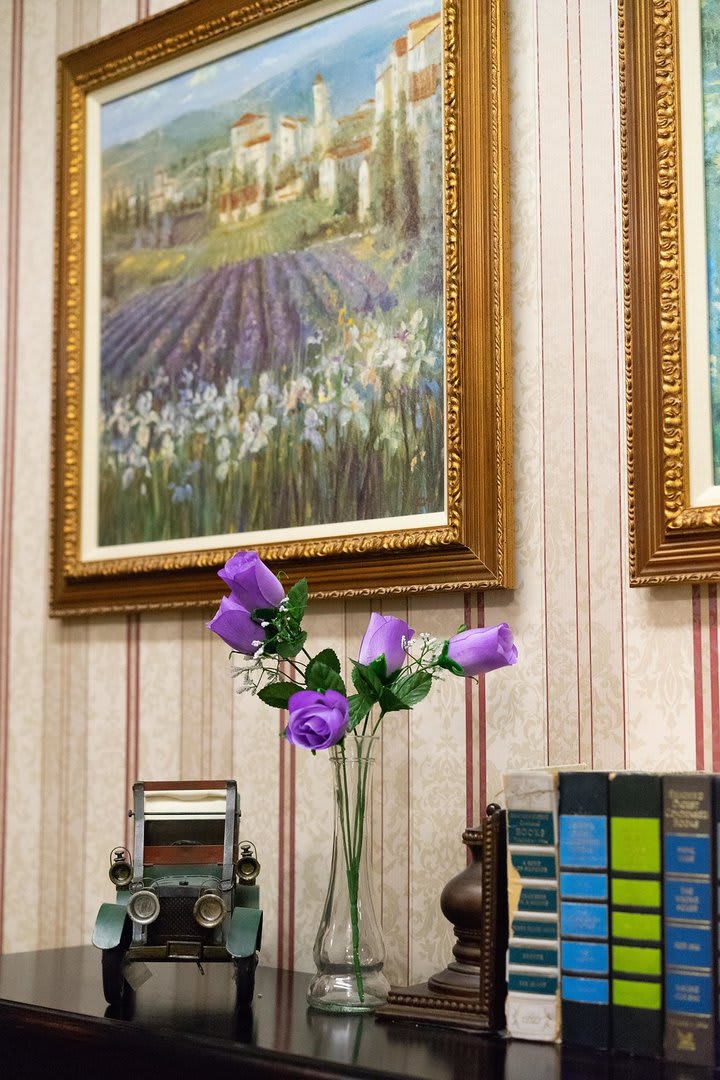 A cozy interior featuring a vase with purple flowers, framed paintings on the wall, and books arranged neatly.