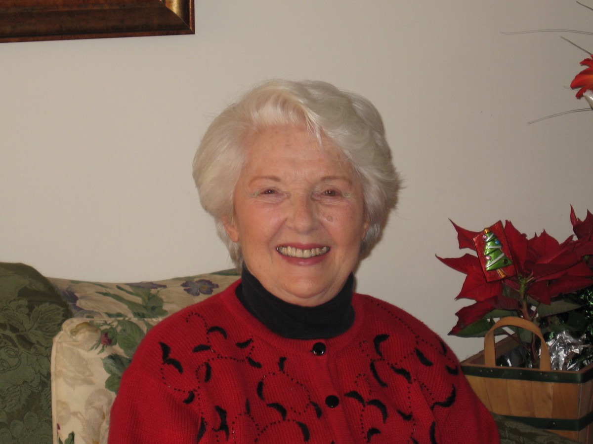 An elderly woman with white hair is smiling indoors, wearing a red sweater.