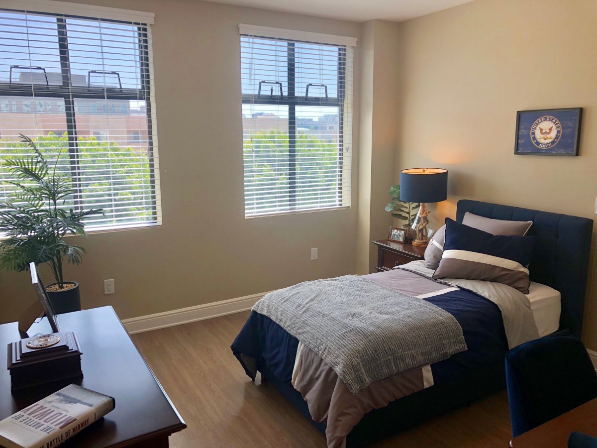 A cozy bedroom with a single bed, nightstand, and large windows with blinds.