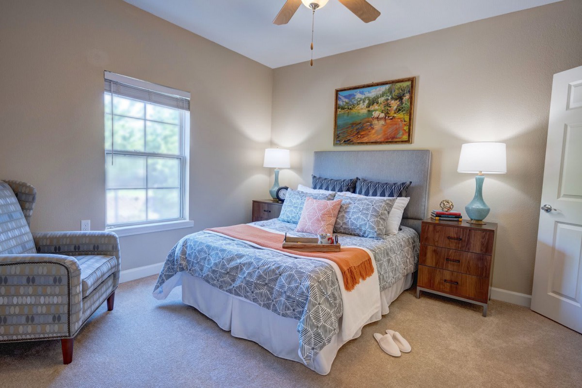 A cozy bedroom with a neatly made bed, soft lighting from bedside lamps, and a large window bringing in natural light.
