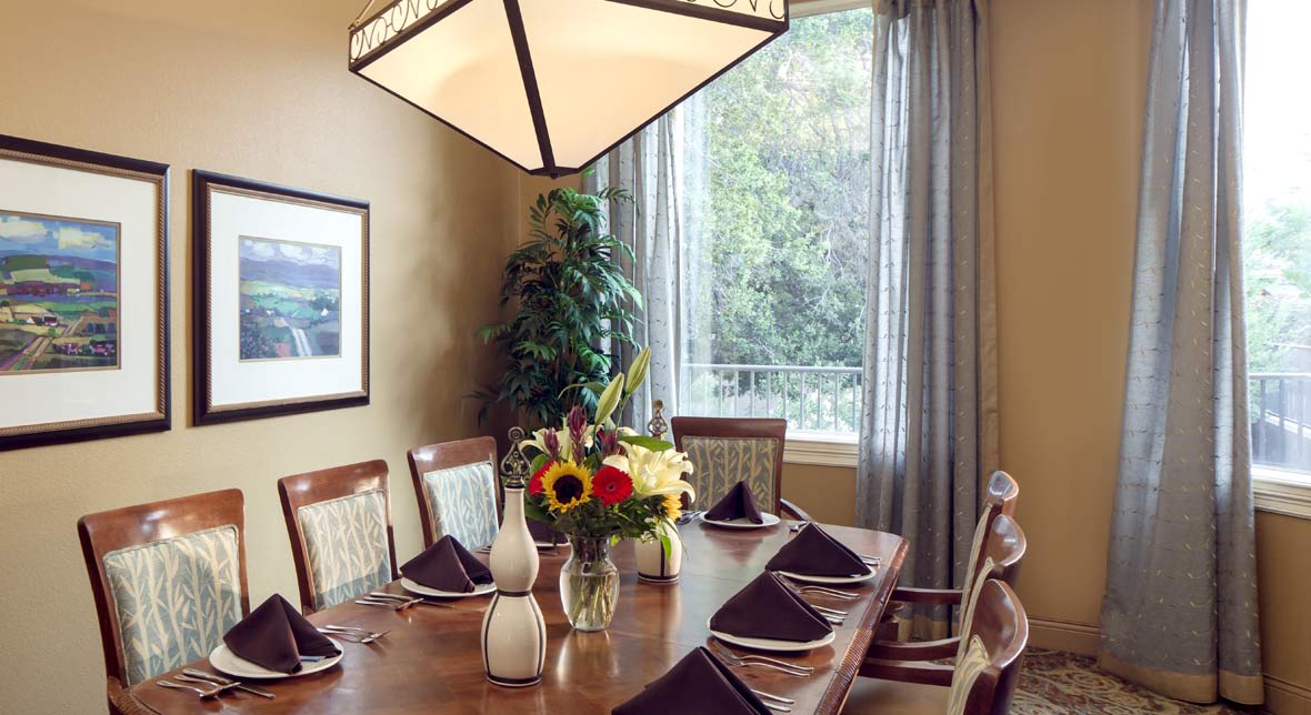 A well-set dining room table with floral centerpiece and elegant napkins, next to a large window with curtains.