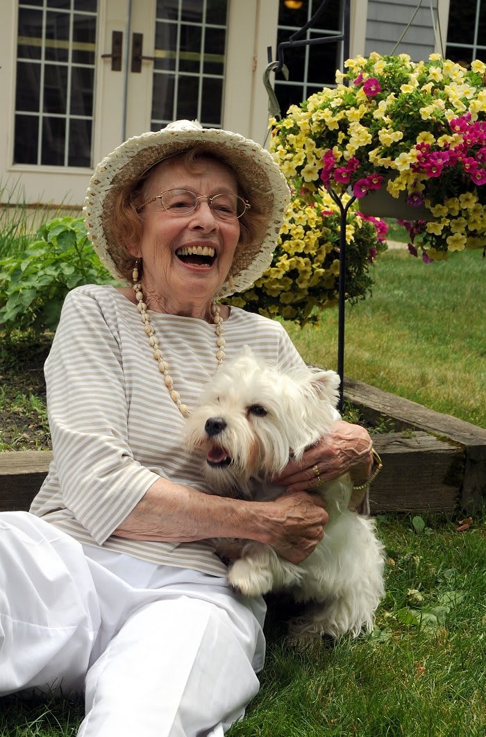 An elderly woman wearing a hat is joyfully holding her dog in a garden surrounded by flowers.