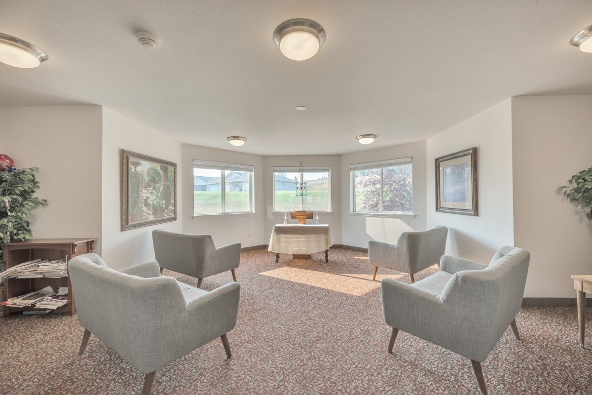 A cozy room with four gray chairs arranged around a central table, surrounded by artwork and windows offering views.