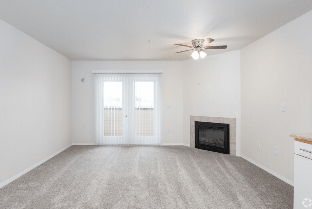 A minimalist living room featuring a fireplace, ceiling fan, and large windows allowing natural light.