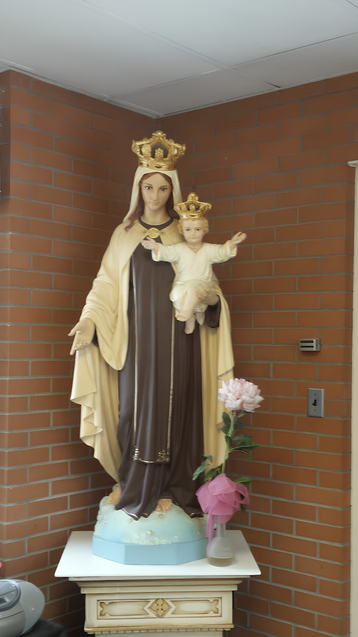 A statue of the Virgin Mary holding a child, adorned with crowns, set against a brick wall with a flower placed nearby.