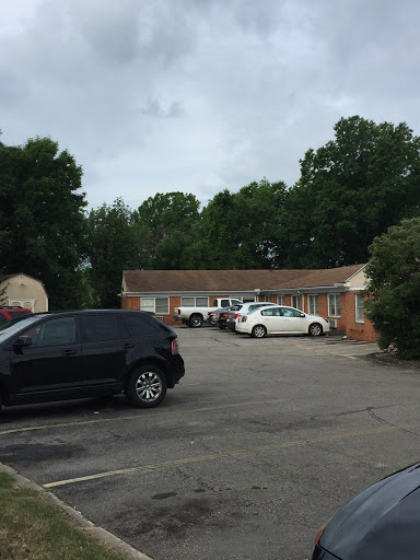 A view of a motel parking lot with several cars parked and surrounded by trees under a cloudy sky.