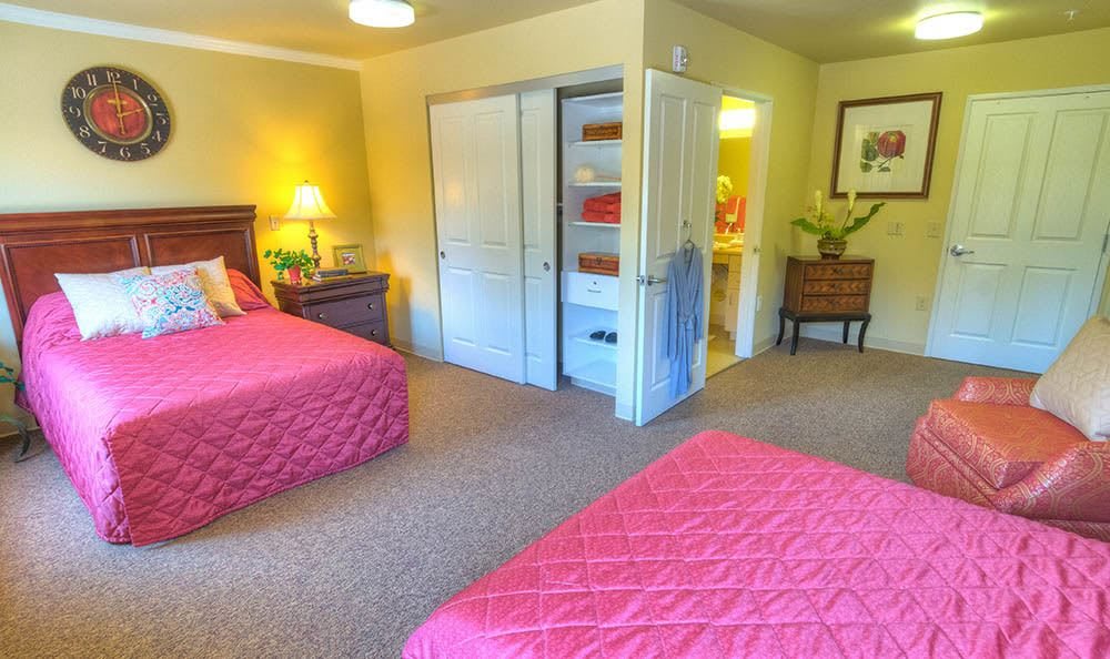 A cozy bedroom featuring two double beds with red quilts, a bedside table and lamp, an open closet, and carpeted flooring.