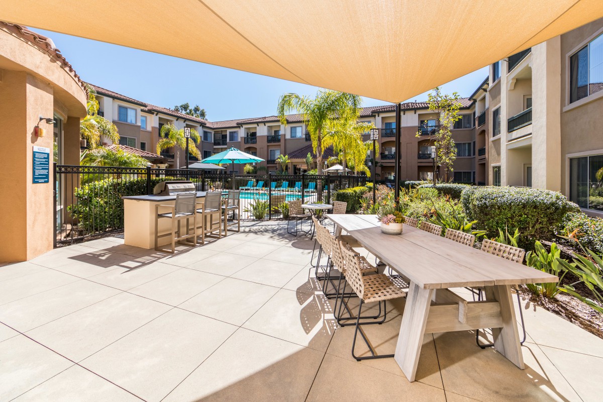 A modern apartment complex features a well-maintained pool area with outdoor seating and palm trees under a shaded patio.