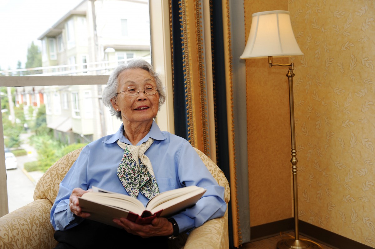 An elderly woman sitting comfortably in a chair, reading a book, with a lamp and window in the background.