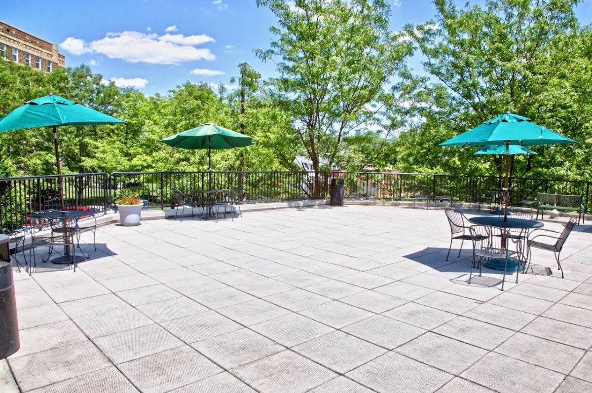 A spacious outdoor patio with tables and umbrellas under a bright, sunny sky surrounded by trees.