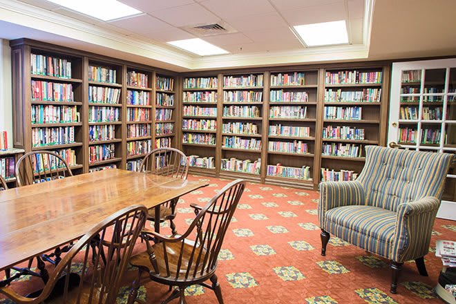 A cozy library room with bookshelves, a wooden table, chairs, and an armchair on a carpeted floor.