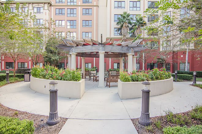 A serene courtyard featuring a pergola with outdoor seating, surrounded by lush landscaping and framed by an apartment building.