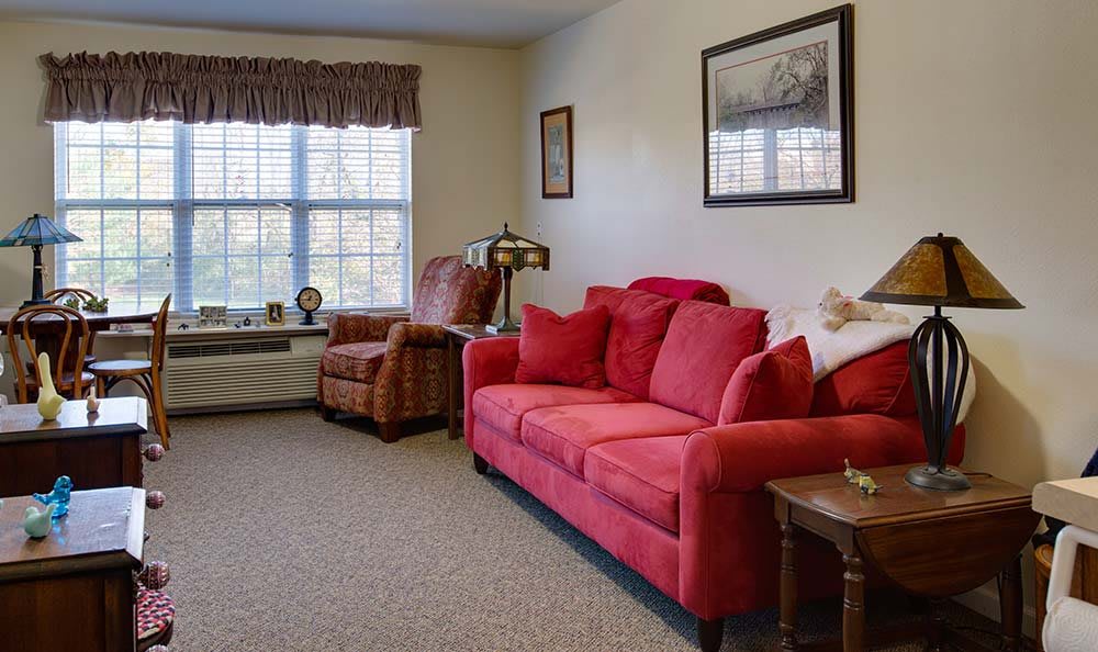 A cozy living room featuring a red sofa and an armchair by a large window, decorated with wall art and a lamp.