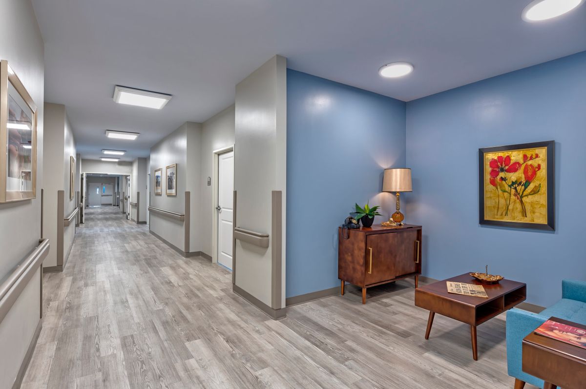 A well-lit hallway in a nursing home with blue walls and wooden flooring, featuring a lamp on a table.
