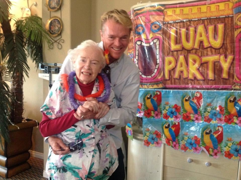 A man and an elderly woman celebrate at a luau-themed party with colorful decorations.