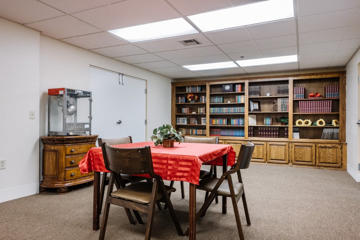 A cozy library room with shelves full of books, a table covered with a red tablecloth, and a popcorn machine in the corner.