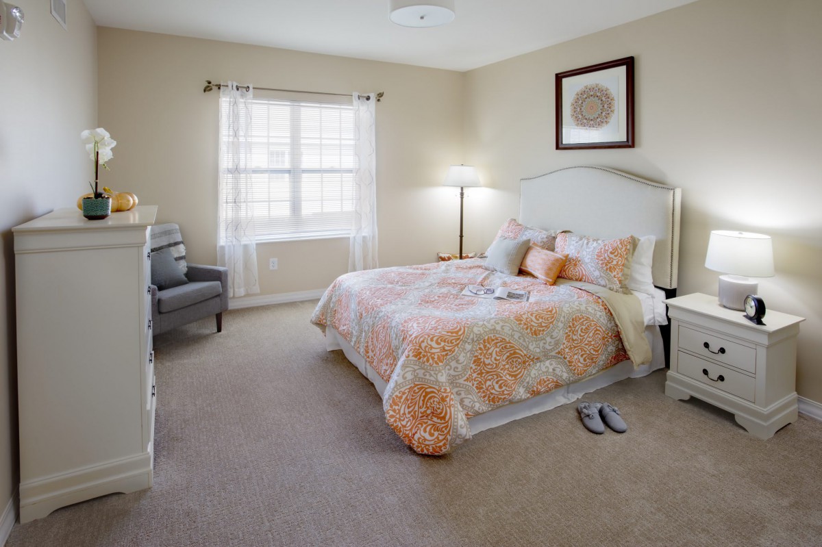 A cozy bedroom with a bed featuring floral bedding, a lamp, and a chair by the window.