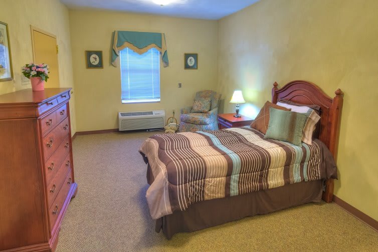 A cozy bedroom interior featuring a bed with colorful bedding, a bedside lamp, and an armchair by the window.