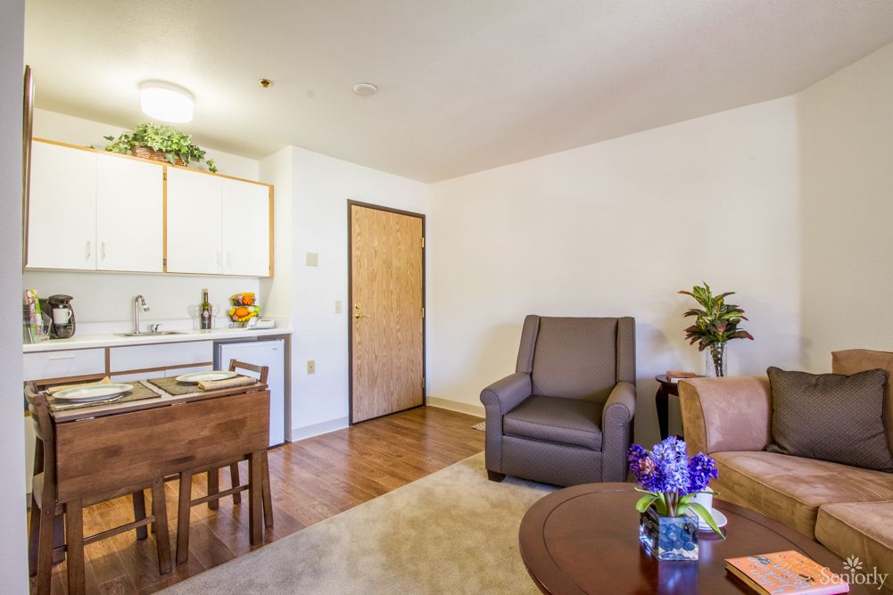 A cozy living room with a kitchenette, featuring a comfortable chair, table with flowers, and wood flooring.
