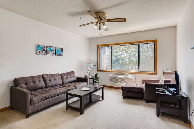 A cozy living room with a brown sofa, coffee table, ceiling fan, and large window.