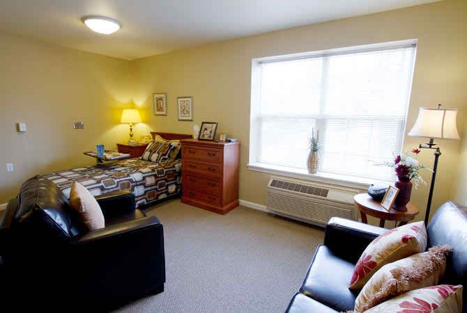 A cozy bedroom featuring a bed, dresser, and sofa with natural light from a large window.