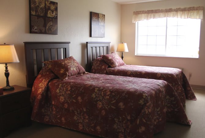 A cozy bedroom with twin beds adorned with floral bedding, a window letting in natural light, and a bedside lamp for evening ambiance.