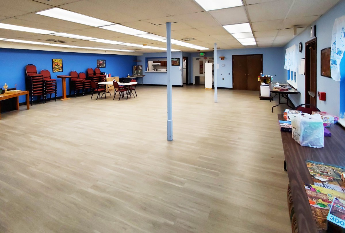 An empty community room with blue walls, featuring tables and chairs, under fluorescent lighting.