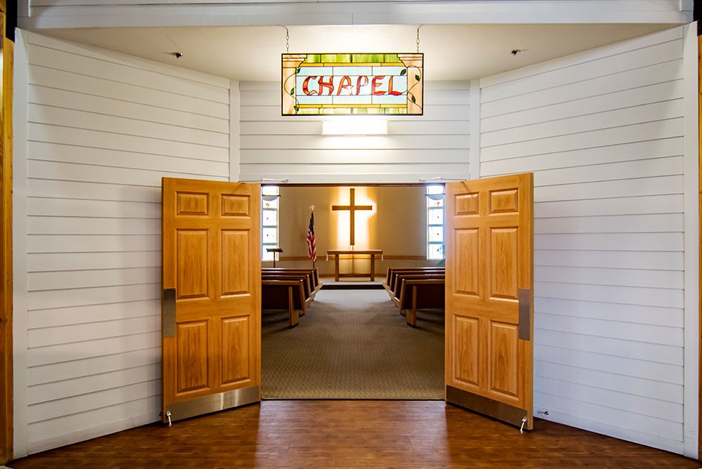 An inviting chapel with wooden doors open, revealing a cross at the front and rows of seating inside.