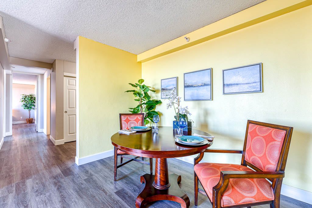 A cozy dining area with a round table and two chairs, set against a yellow wall adorned with artwork and complemented by plant decor.