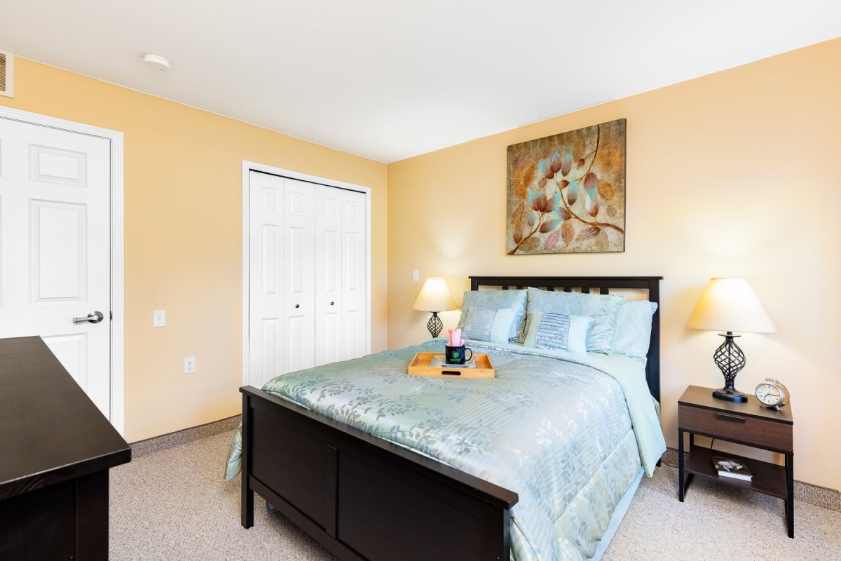 A cozy bedroom with a neatly made bed, adorned with cushions and a decorative tray, complemented by warm lighting and artwork on the wall.