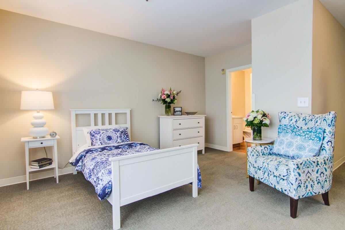 A cozy bedroom with a single bed, decorated with floral bedding, an armchair, and a dresser topped with fresh flowers.