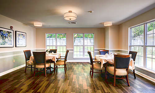 A cozy dining room with neatly arranged tables and chairs, featuring large windows and elegant lighting.