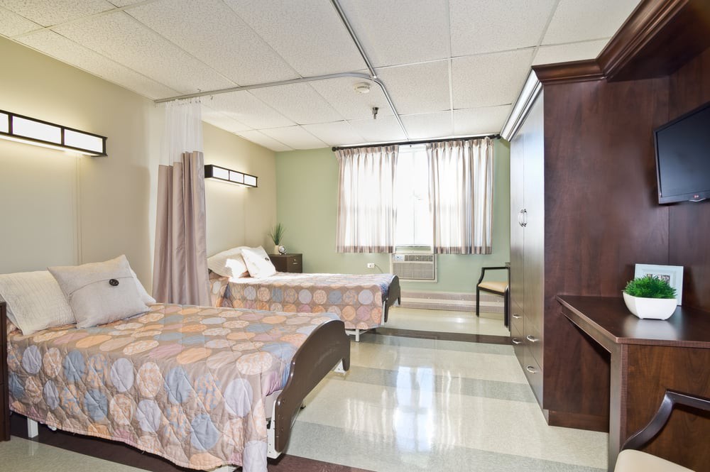 A hospital room with two beds, separated by a curtain, and a window providing natural light.
