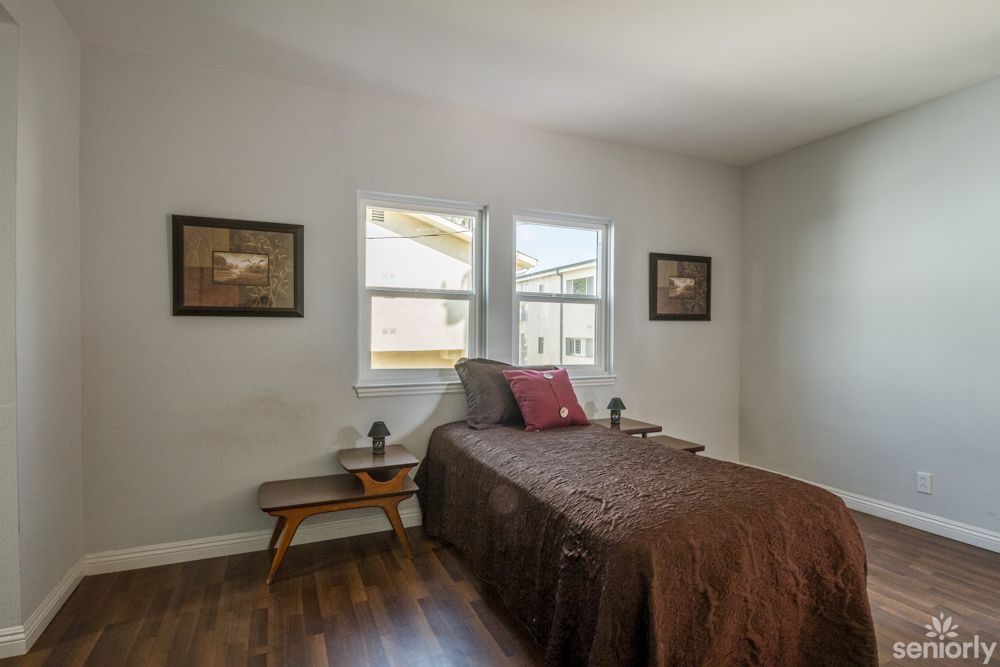 A cozy bedroom with a single bed, artwork on the walls, and natural light from two windows.