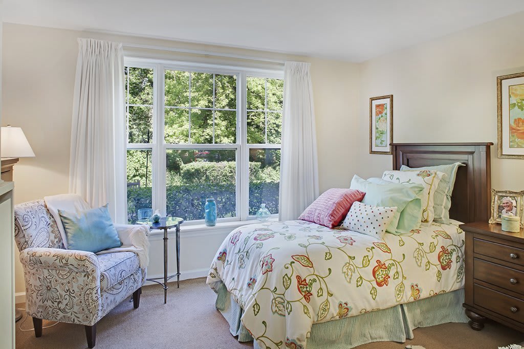 A cozy bedroom with a large window allowing natural light to fill the space, featuring a bed with floral-patterned bedding and an armchair.