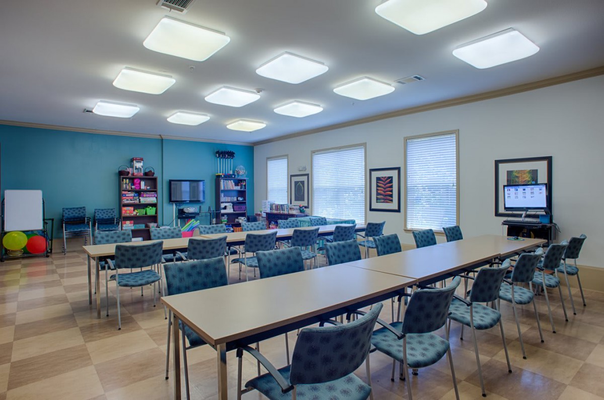 An empty classroom with tables and chairs arranged in rows, illuminated by ceiling lights and featuring bookshelves along the wall.