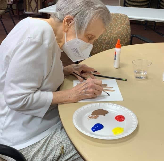 An elderly person wearing a face mask is painting at a table with art supplies.