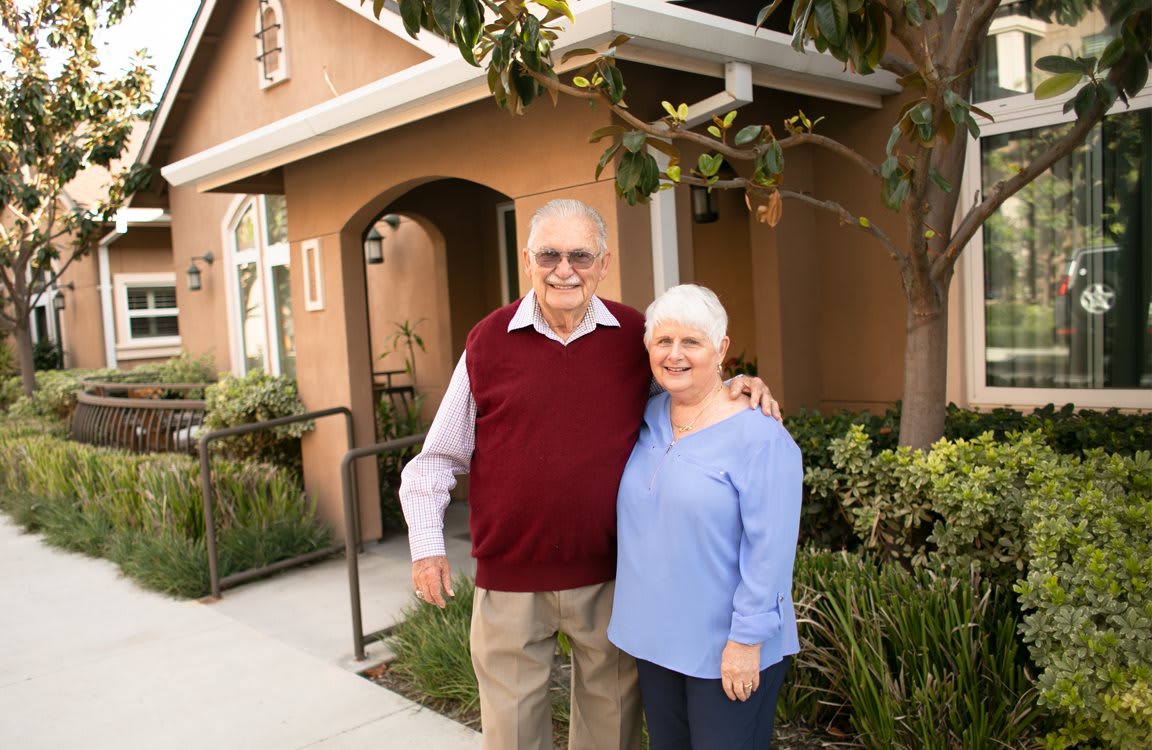 An elderly couple smiling and standing together in front of their house.