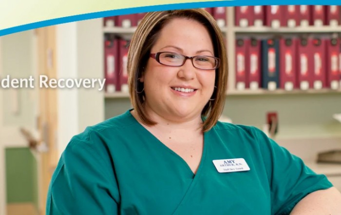 A female healthcare professional wearing glasses and a uniform stands in front of a bookshelf.